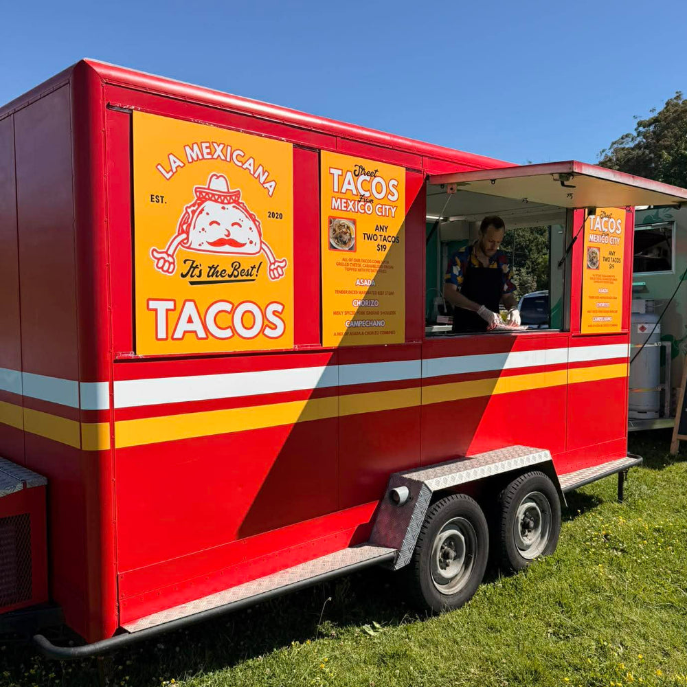 Red taco food truck with 'La Mexicana' branding on a grassy area.